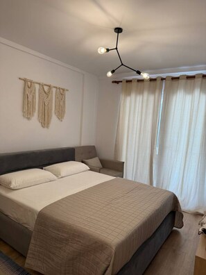 Quarto com cama grande, cabeceira, mesa de cabeceira, luminária e janela com cortinas.