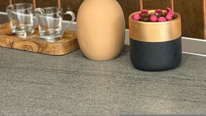 Un jarrón beige y un recipiente negro y dorado con flores rosadas sobre una bandeja de madera.
