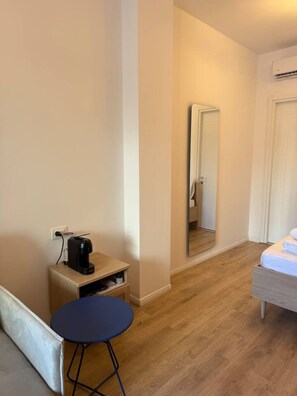 Ein kleiner, ordentlicher Raum mit einem Bett, einem Nachttisch mit Kaffeemaschine, einem kleinen Hocker und einem Spiegel an der Wand.