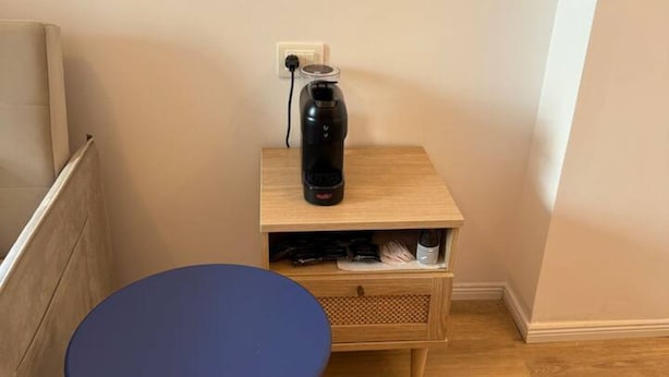 Una habitación pequeña con una mesa azul, una mesita de noche de madera y un objeto negro encima, y un enchufe de pared.