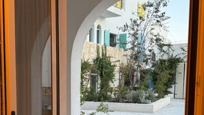 Una puerta abierta que da a un patio con suelo de piedra, plantas en macetas y vistas de edificios con contraventanas azules.