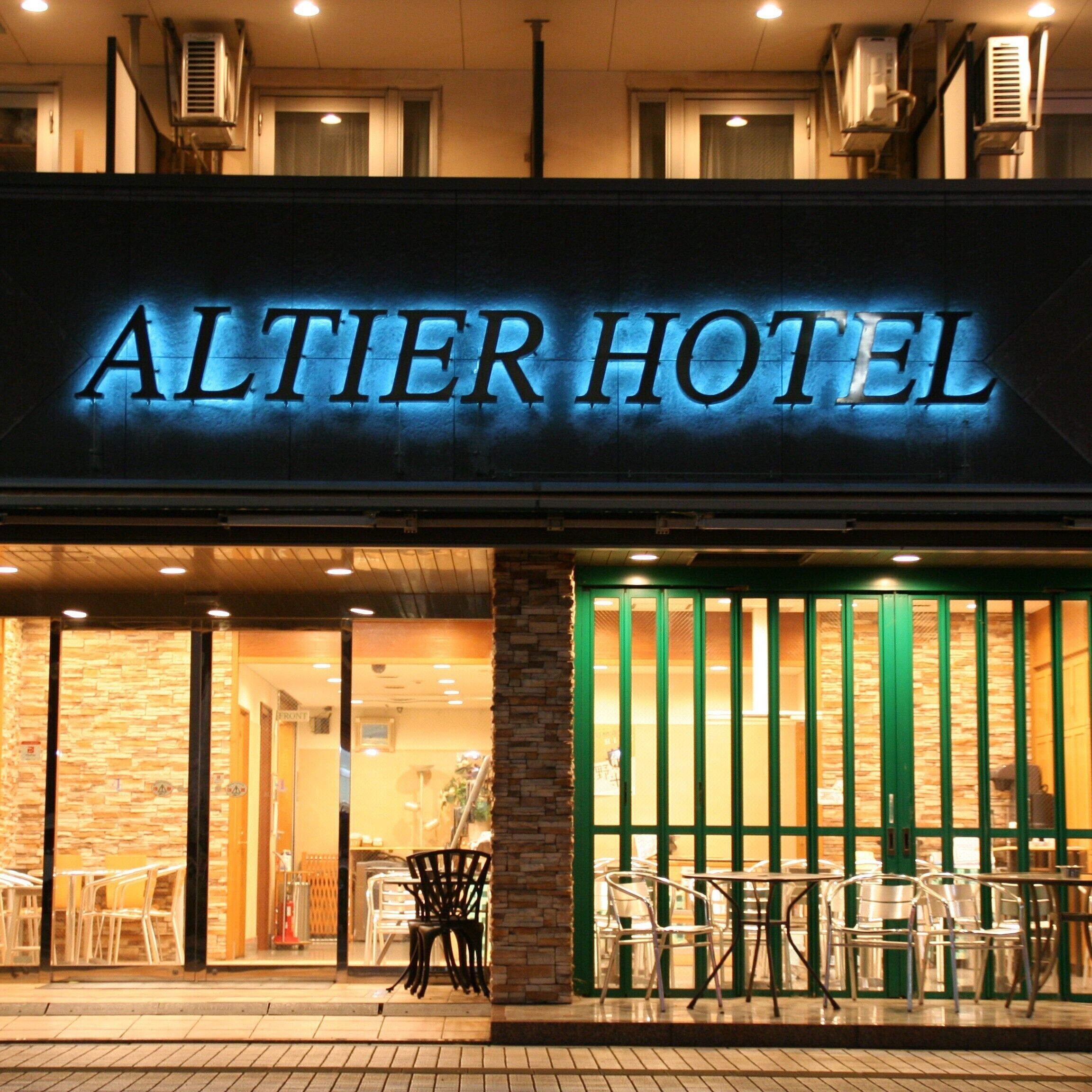 The exterior of the Altier Hotel with a prominent illuminated sign, large windows, and outdoor seating area.