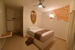 A compact room with a bed, a small table, a shelf, a ceiling fan, and a decorative wall pattern. - Hotel Aldaria Bacalar (Bacalar)