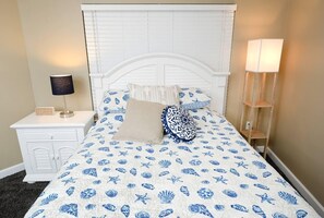 A bedroom with a white headboard, a bed with a blue and white seashell pattern bedspread, two pillows, a bedside table with a lamp, and a nightstand with a digital clock. - Shores of Panama 530 (Panama City Beach)