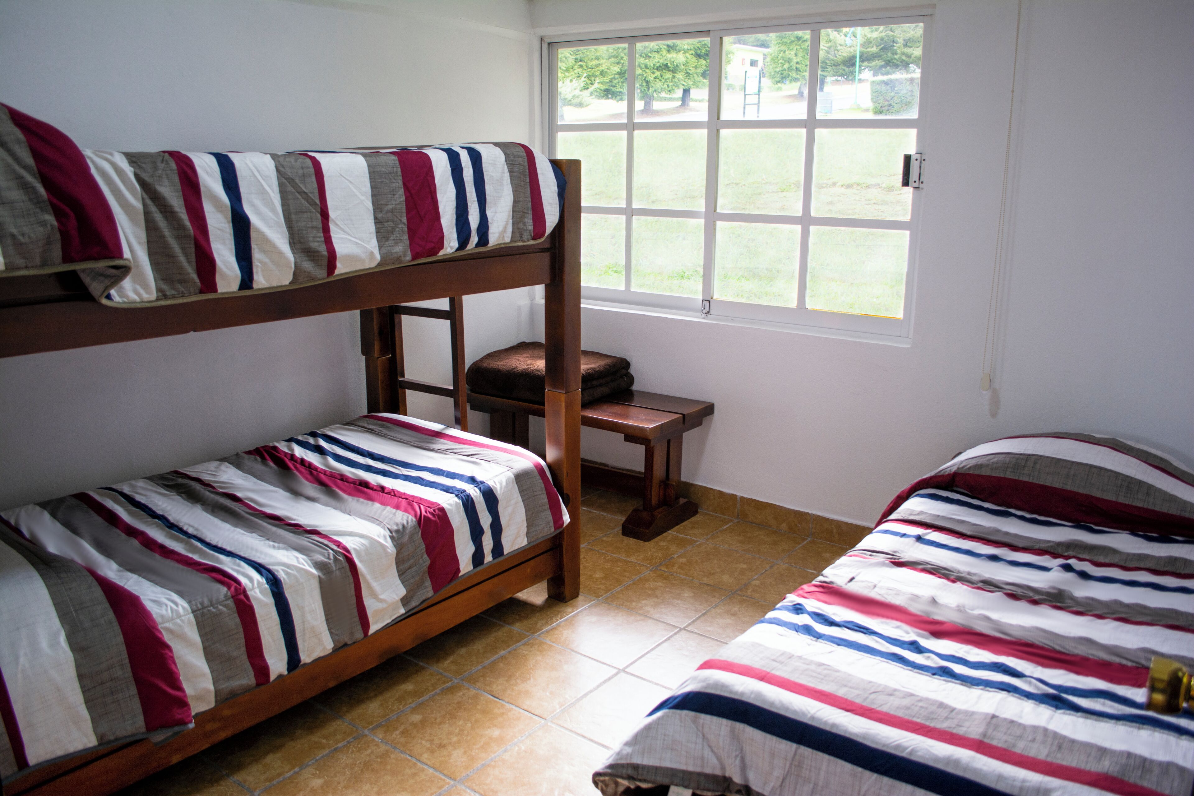 Habitación con literas, vista al exterior por una ventana, una pequeña mesa de madera y ropa de cama a rayas.