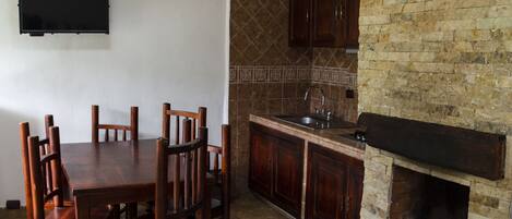 A dining area with a wooden table and chairs, a kitchen with a brick wall and tiled backsplash, and a flat-screen TV mounted on the wall.