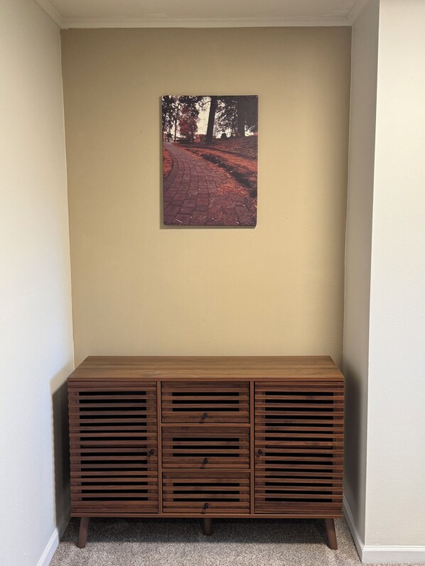 Una habitación de esquina con una pared color beige, un banco de madera con cajones y un cuadro con una imagen de un sendero.