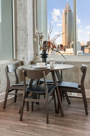 Mesa redonda de comedor con cuatro sillas, un jarrón con plantas secas, un cuenco blanco y un vaso de agua. La habitación tiene una pared de concreto y un ventanal con vistas a la ciudad.