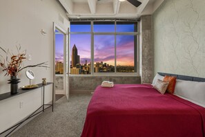 A bedroom with a city view, a bed with a red cover, a bedside table with a mirror, and a vase with dried branches. - Downtown ATL High-Rise | Kings, Views & Pool (Atlanta)
