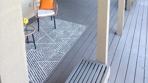 Una terraza techada al aire libre con una silla y una mesita, una ventana con persianas y baranda de madera.