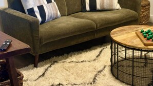 Una sala de estar con un sofá verde, almohadones a rayas, una mesa redonda de centro con manzanas, una alfombra mullida y una obra de arte floral en la pared.