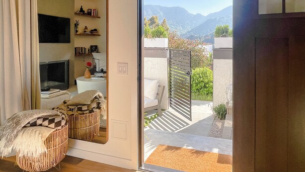 A room with a large mirror, a television, and a wicker chair. There is a sliding door leading to an outdoor area with a view of mountains. - 2BD/2BA Palisades Townhouse Monthly Rental (Los Angeles)