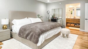 A bedroom with a large bed, bedside table, lamp, dresser, mirror, and a small stool. - 2BD/2BA Palisades Townhouse Monthly Rental (Los Angeles)