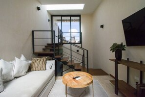 Un salon moderne avec un canapé, une table basse ronde en bois, un escalier doté de rampes en verre et une vue sur l’extérieur.