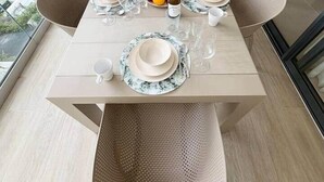 Une table à manger est dressée avec des assiettes, des verres et une bouteille, entourée de chaises en osier, dans une pièce dotée d’une grande fenêtre.