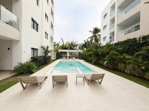 Uma área de piscina moderna com três cadeiras de descanso e uma mesa de bilhar, cercada por vegetação exuberante e fachada de um edifício branco.