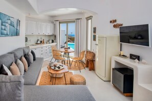 Ein modernes, helles Wohnzimmer mit grauem Sofa, Korbstühlen und einem Essbereich mit Tisch und Stühlen. An der Wand ist ein Flachbildfernseher angebracht, und im Hintergrund ist eine Küche mit weißen Schränken zu sehen.