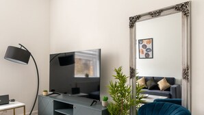 A modern living room with a flat-screen TV, a blue armchair, a floor lamp, a grey entertainment unit, and a large ornate mirror. - Modern Flat in Waterloo (London)