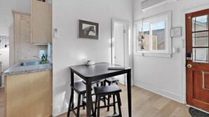 Une cuisine avec un meuble en bois, un évier et une table avec deux tabourets. Il y a un tableau encadré au mur et une porte avec une fenêtre.