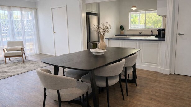 A dining area with a dark wooden table and four upholstered chairs, a vase with flowers, and a kitchen in the background.