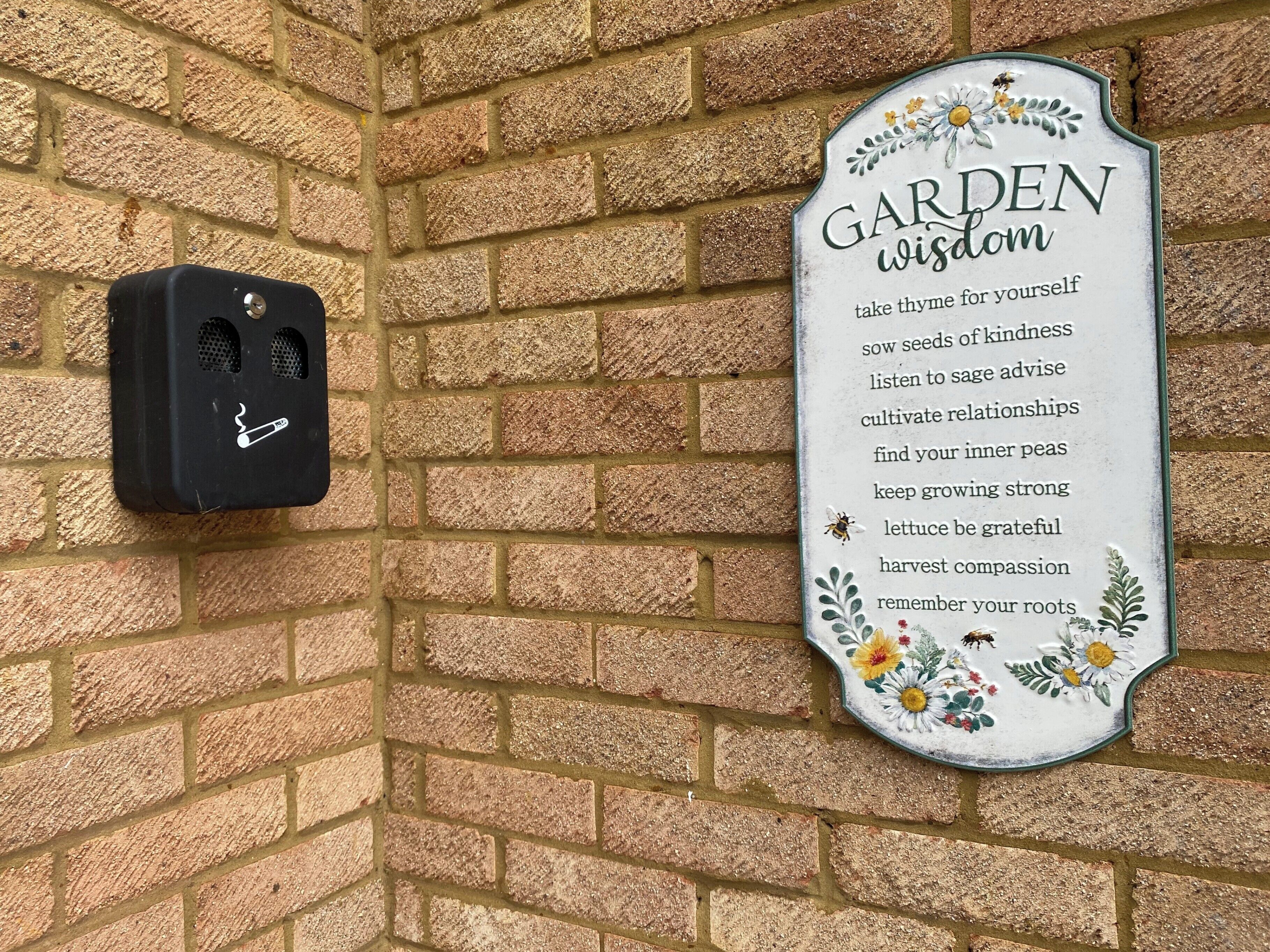 A brick wall with a sign that reads "GARDEN WISDOM" and a smoke detector mounted on it.