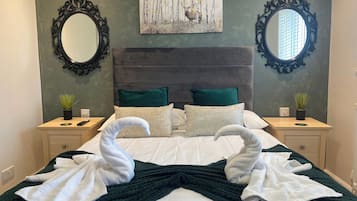 Uma cama bem arrumada com duas toalhas modeladas em forma de cisnes, um cobertor verde escuro e duas almofadas decorativas.