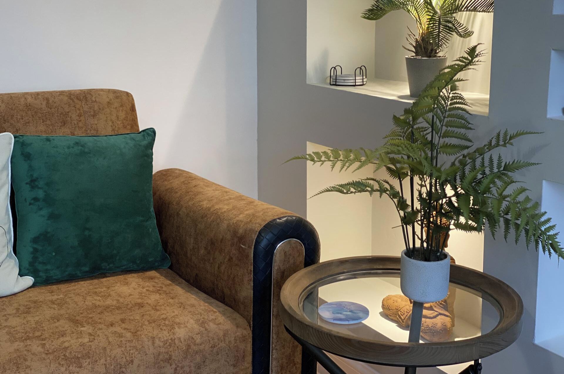 A living room with a brown sofa, a green pillow, a round glass table with a plant, and a small shelf with a potted plant and a decorative item.