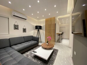 Ein modernes Wohnzimmer mit einer grauen Eckcouch, einem weißen Couchtisch und einer schwarzen Stehlampe. Es gibt einen Essbereich mit einem Holztisch und Stühlen, und im Hintergrund ist eine Küche zu sehen.