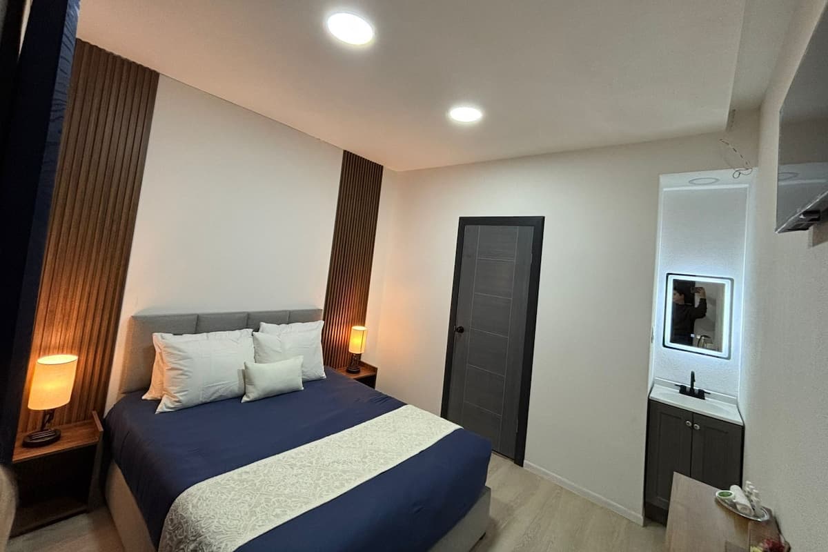 A neatly arranged bedroom with a bed, bedside lamps, a door, a mirror, and a small cabinet.