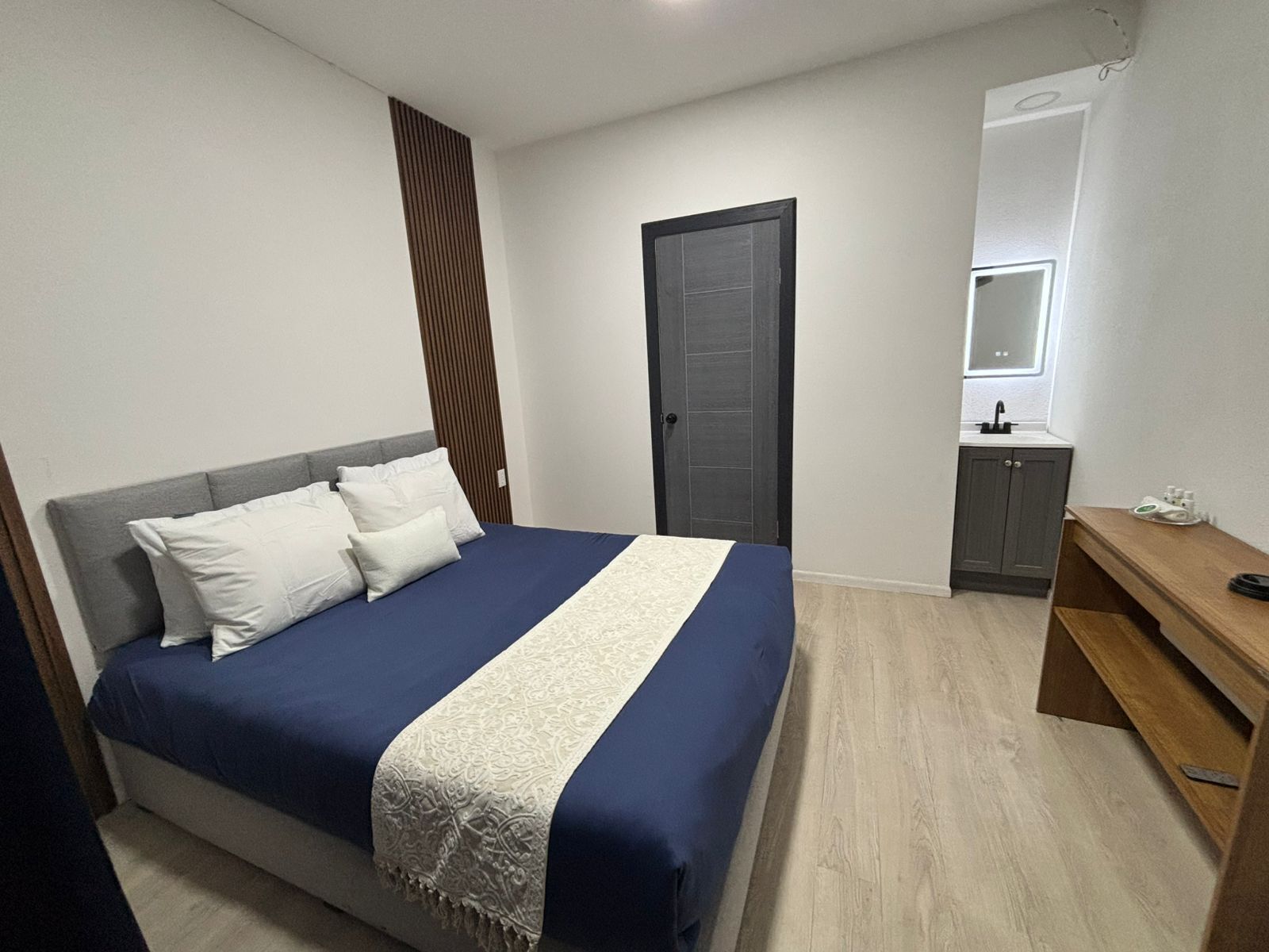 A bedroom with a bed, a bedside table, a wardrobe, and a bathroom with a mirror and sink.