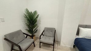 A small, modern room with two grey armchairs, a small potted plant, and a bed with a blue blanket.