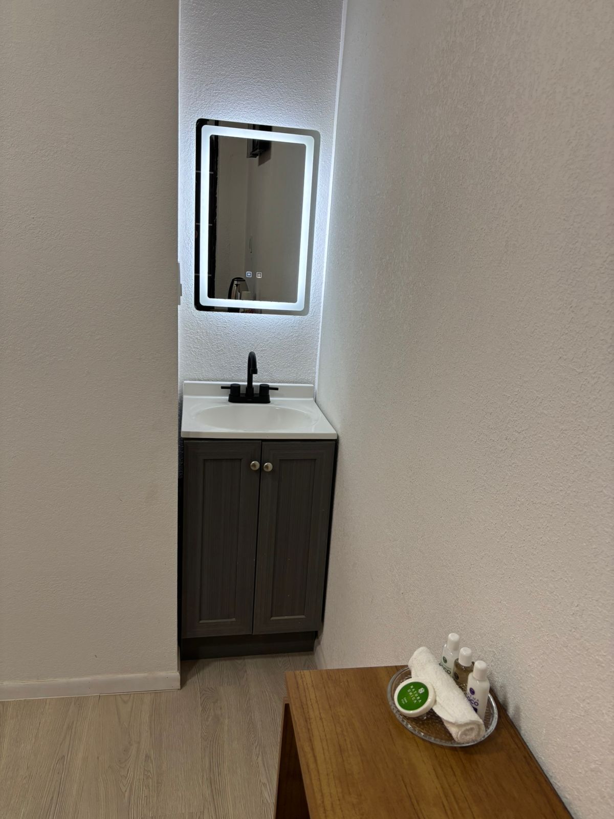 Un bagno con un lavandino bianco, mobili scuri, uno specchio con luce integrata e un tavolino con articoli da toilette.