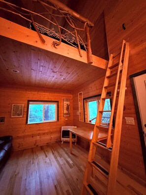 A cabin interior with a loft bed, a ladder, a heater, and two windows with a view of the forest.