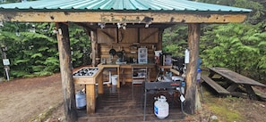 A rustic wooden structure with a gas stove, sink, and various cooking utensils.