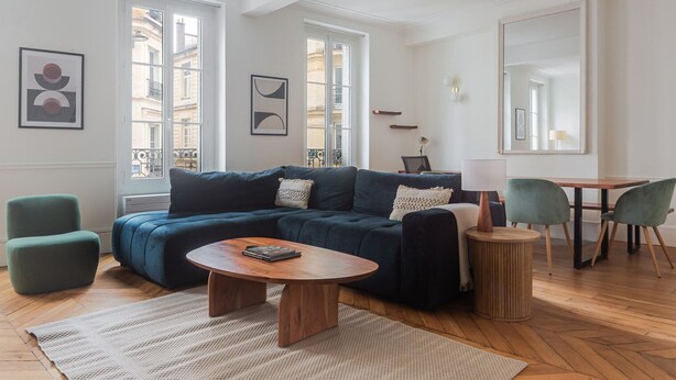 A modern living room with a dark blue sectional sofa, a round coffee table, and a dining area with a wooden table and two chairs. - Blueground | Épinettes, view, nr Parc Monceau (Paris)