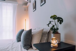 Una camera da letto ordinata con un letto, cuscini, un comodino con una pianta e una lampada sferica, e una cornice appesa al muro.