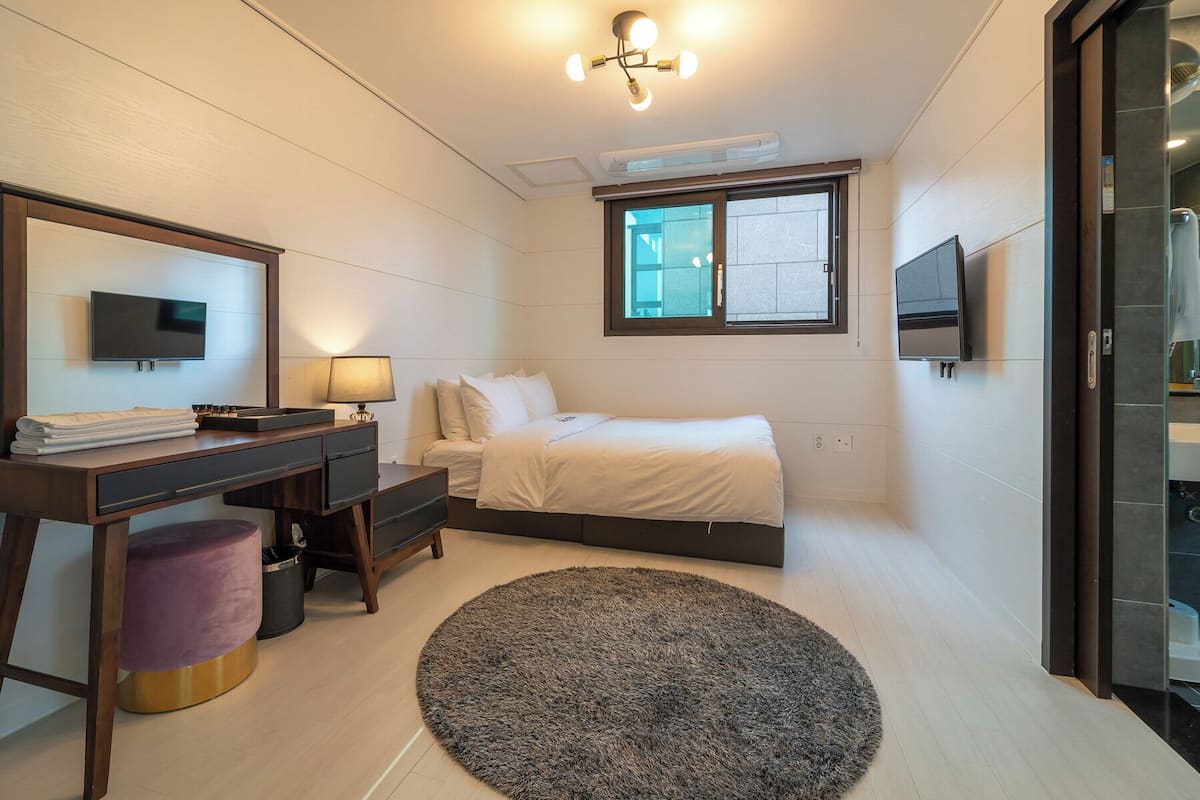 A bedroom with a bed, a desk, a chair, a TV, and a window.