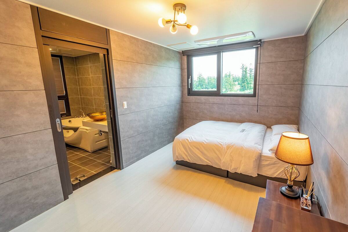 A modern bedroom with a bed, bedside lamp, and a window with a view of trees.