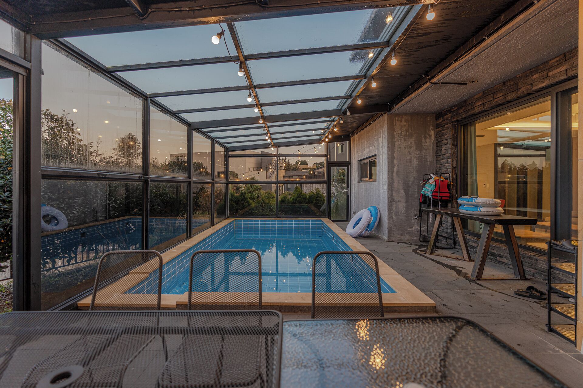 Une piscine intérieure, entourée de parois en verre, offrant une vue sur les environs, ainsi qu’un espace salon couvert et une table sur laquelle sont disposés divers objets.