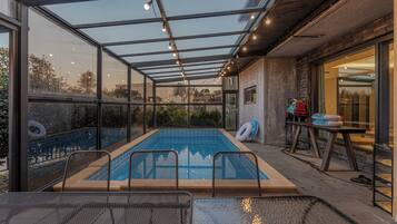 Une piscine intérieure, entourée de parois en verre, offrant une vue sur les environs, ainsi qu’un espace salon couvert et une table sur laquelle sont disposés divers objets.