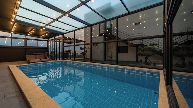An indoor swimming pool with a glass ceiling, a sun terrace with seating, and a view of the surrounding landscape. - Jeju Calma (Jeju City)