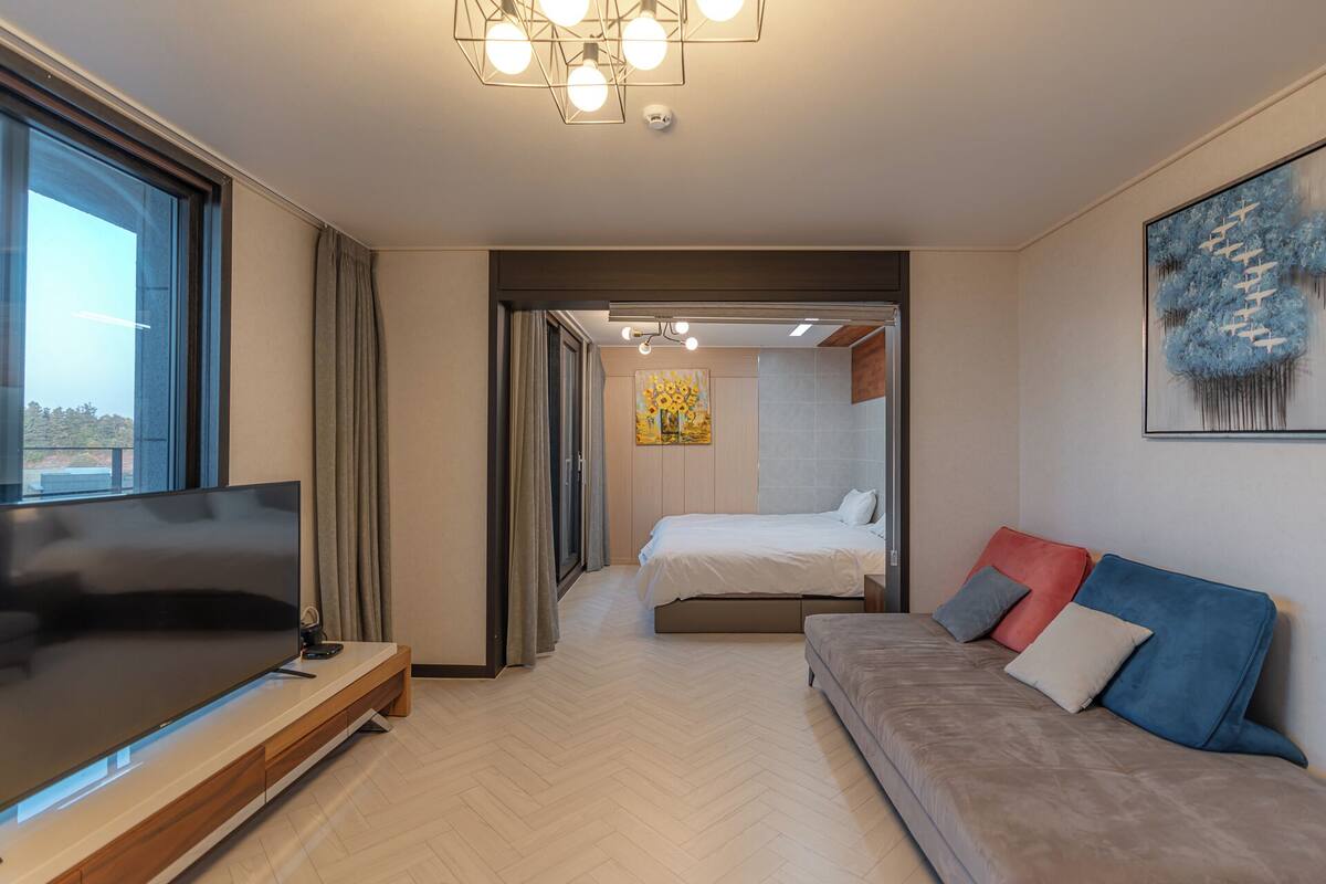 A modern hotel room with a large flat-screen TV, a sofa with cushions, a bed, and a painting on the wall.