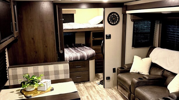 Een compact interieur van een camper met een kitchenette, een eethoek, een zithoek met een bank en een slaapbank, en een kleine tafel met een plantje.