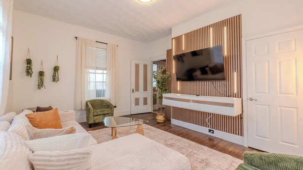 Un salon moderne avec un canapé, un fauteuil vert, une table basse en verre et un téléviseur à écran plat fixé sur un mur recouvert de panneaux en bois.