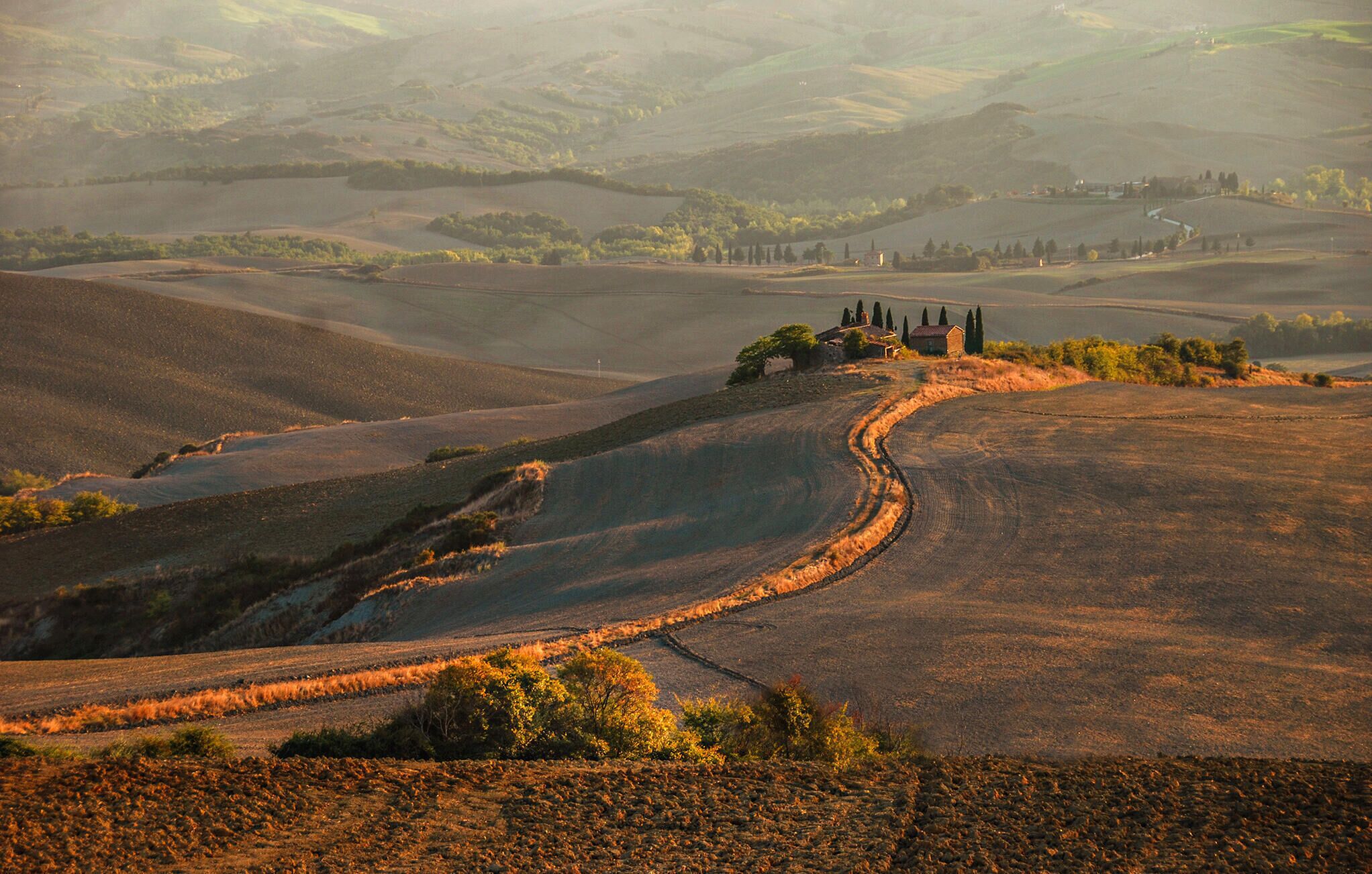 Un paysage rural caractérisé par des collines douces, un sentier sinueux et un petit bâtiment surmonté d’un toit rouge.