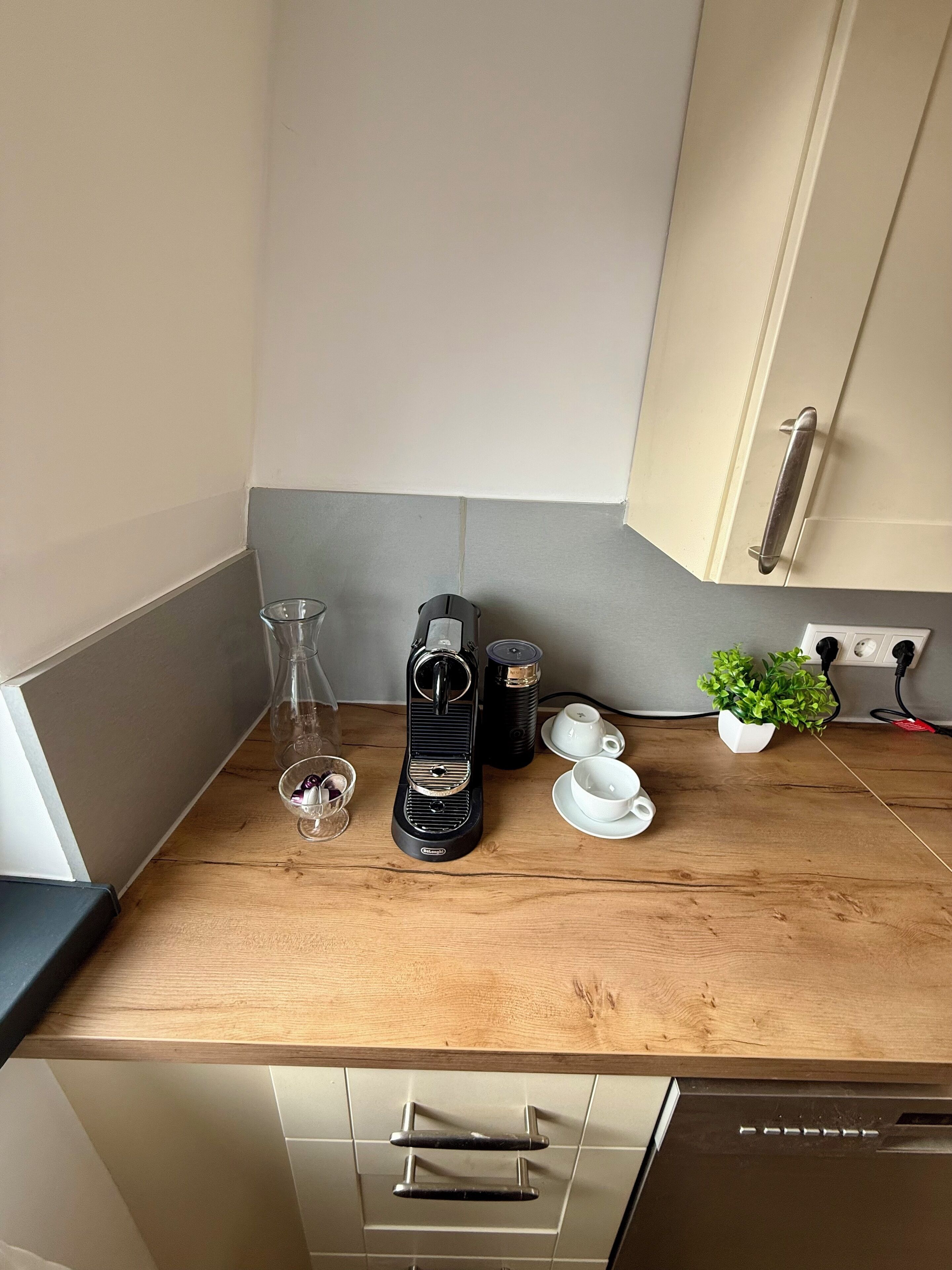 A kitchen counter with a coffee maker, two cups, a glass, and a small plant.