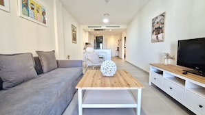 A modern living room with a grey sofa, a wooden coffee table with a decorative sphere, a TV on a wooden stand, and a dining area in the background. - SR Calanova Golf 2BDR with private garden (Mijas)