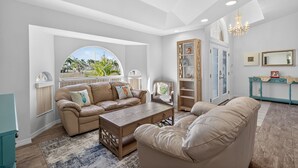 Een ruime woonkamer met een hoge plafond, een groot raam met uitzicht op palmbomen, een beigekleurige zithoek, een houten salontafel en een turquoise console met decoratieve objecten.