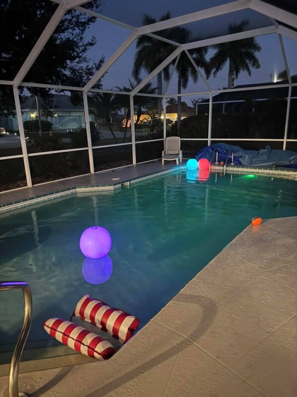 A swimming pool with a slide and inflatable toys, surrounded by a covered area with a view of palm trees and a building in the background. - Fin & Tonic~Heated Pool~Sleeps 12~Elegant Cape Coral Retreat (Cape Coral)