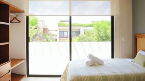 A bedroom with a large window, a bed with a rolled towel, wooden shelves, and a view of greenery and buildings outside. - 🌴 Stylish 3-Bedroom Condo w/ Pool in Aldea Zama - Perfect for Families & Groups (Tulum)